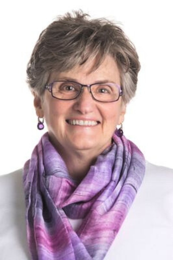 Linda Yetman smiling at the camera with a white background, short greyish hair, and a pink and purple scarf on.